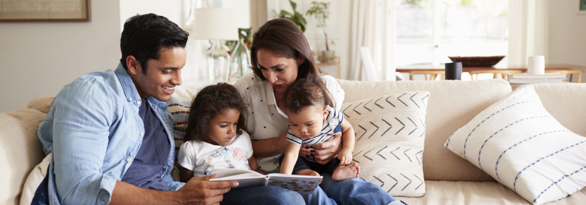 Hispanic couple sitting on the sofa reading a book at home with their baby son and young daughter