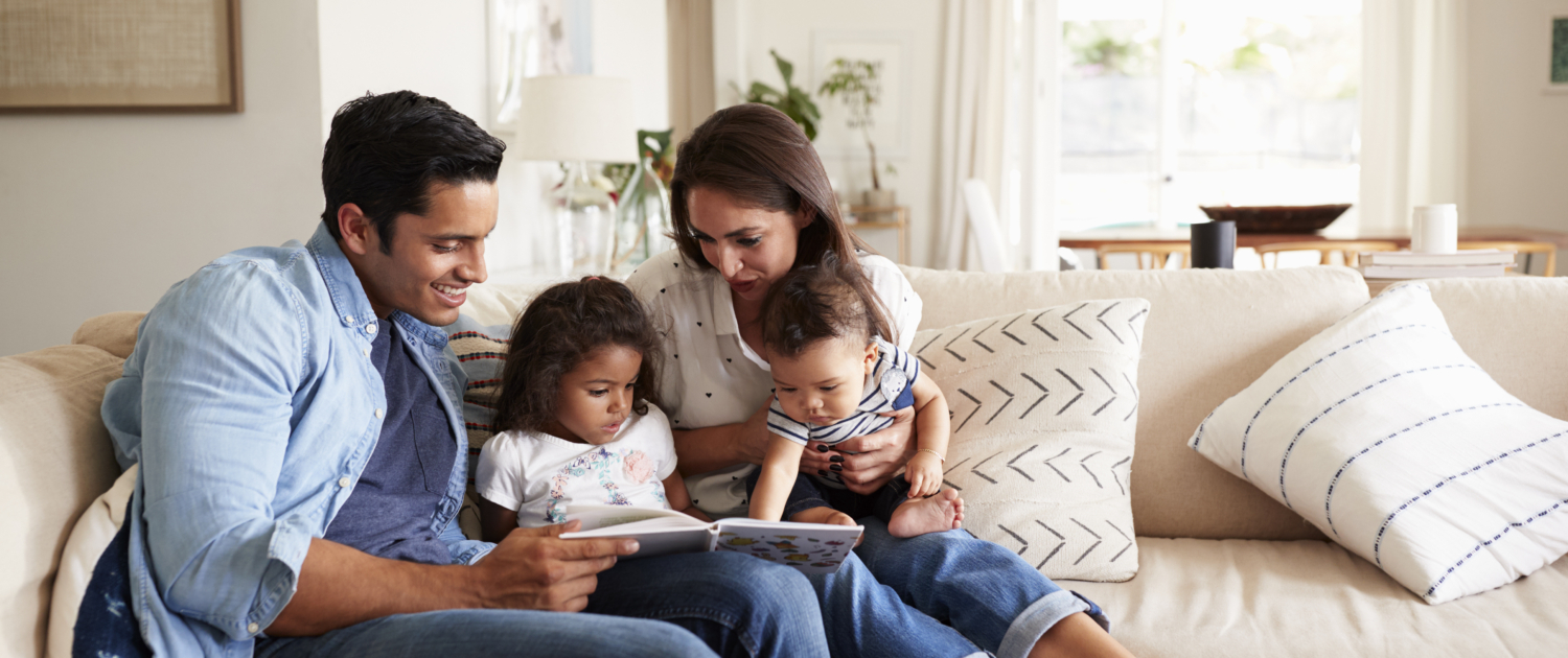 Hispanic couple sitting on the sofa reading a book at home with their baby son and young daughter